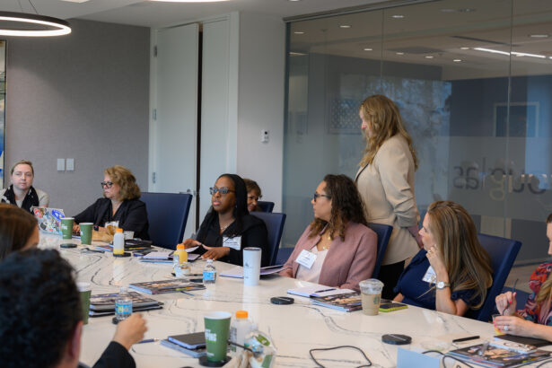 A group of women sit and talk around a conference table in a modern office. Laptops, notebooks, coffee cups, and water bottles are on the table. One woman stands while others listen and engage in discussion. South Florida Business & Wealth
