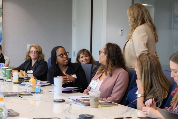 A group of women sit around a conference table in discussion. One woman stands and speaks to another seated woman, while others listen and take notes. Drinks and papers are on the table. South Florida Business & Wealth