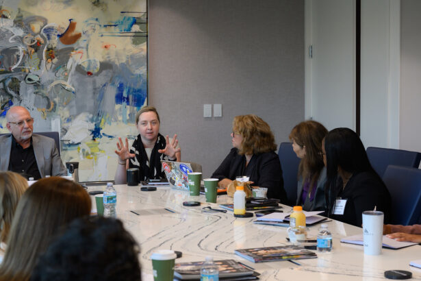 A group of people sits around a conference table in a modern office, engaged in discussion. One woman speaks with expressive hand gestures. Books, drinks, and notebooks are on the table; abstract art is on the wall behind them. South Florida Business & Wealth