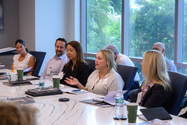 A group of professionals sit around a conference table in a modern office, engaged in discussion. One woman speaks while others listen attentively. Papers, notebooks, and drinks are on the table, with large windows in the background. South Florida Business & Wealth