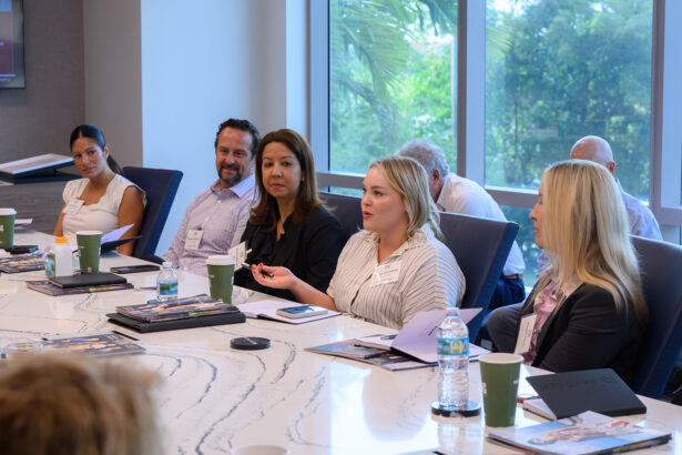 A group of professionals sit around a conference table engaged in discussion. Laptops, notebooks, and drinks are on the table, and large windows behind them let in natural light with a view of greenery. South Florida Business & Wealth