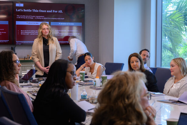 A group of people sit around a conference table in a meeting room, listening to a standing woman. A screen behind her displays a presentation titled "Let's Settle This Once and For All." Daylight streams in through large windows. South Florida Business & Wealth