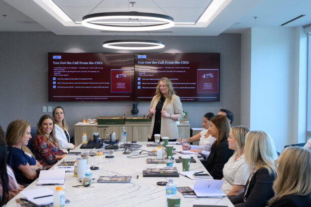 A woman stands and speaks to a group of people seated around a conference table, with notebooks, laptops, and drinks. Two large screens behind her display a presentation with red backgrounds and white text. South Florida Business & Wealth