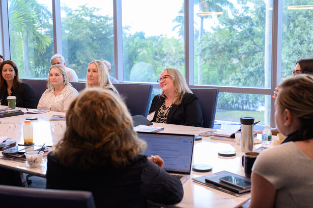 A group of people sit around a conference table in a bright meeting room with large windows and trees visible outside, engaged in discussion with laptops, notebooks, and drinks on the table. South Florida Business & Wealth