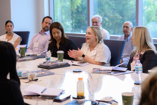 A group of people sit around a conference table in a bright meeting room, engaged in discussion. One woman in the center is speaking, while others listen attentively. There are papers, drinks, and folders on the table. South Florida Business & Wealth