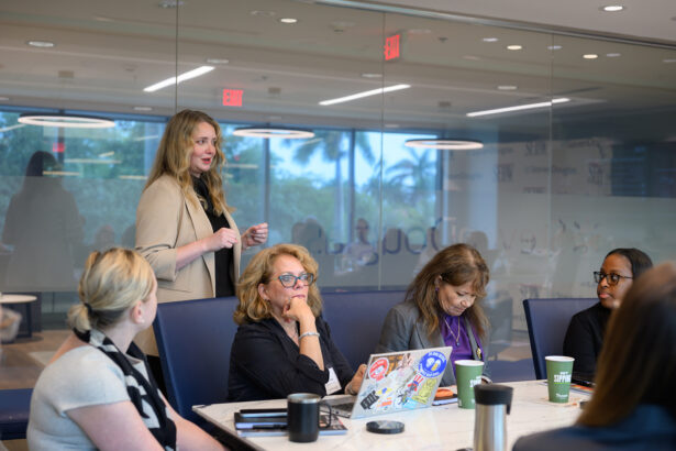 A woman stands and speaks to four seated women around a conference table in a modern office with glass walls. Laptops, coffee cups, and notebooks are on the table. The atmosphere appears collaborative. South Florida Business & Wealth