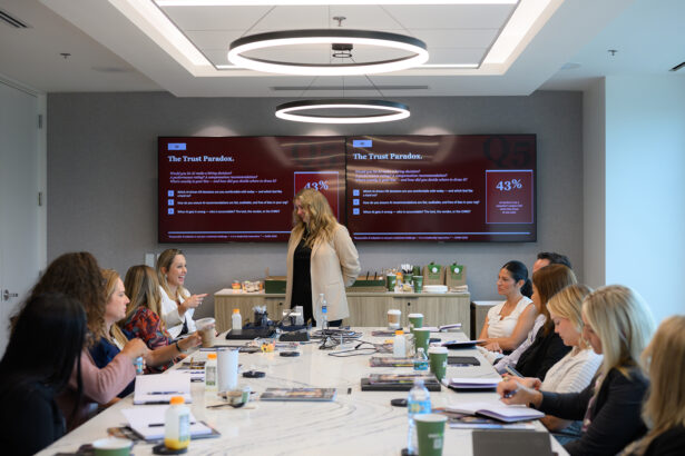 A group of professionals sit around a conference table as a woman stands and speaks in front of two large screens displaying presentation slides titled "The Trust Paradox." Laptops, notebooks, and drinks are on the table. South Florida Business & Wealth