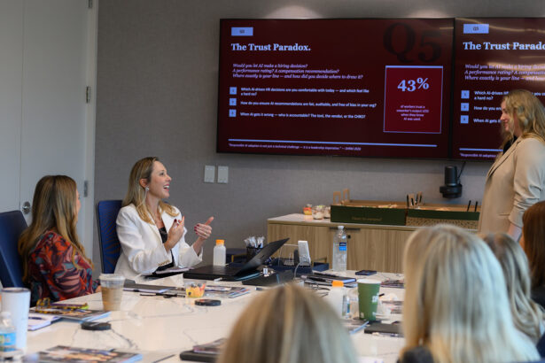 A group of women sits around a conference table engaged in discussion as one woman stands and presents a slide titled "The Trust Paradox" on a large screen, showing a statistic and questions about trust and recommendations. South Florida Business & Wealth