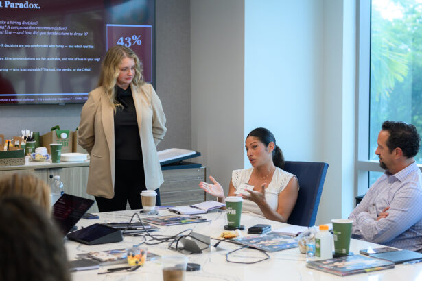 Three people in a meeting room discuss around a table with laptops, papers, and drinks. One woman stands by a screen displaying a chart, while another woman gestures as she speaks. A man listens attentively. South Florida Business & Wealth