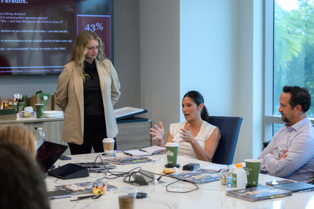 Three people sit around a conference table with laptops, papers, and coffee cups, while one woman stands and another gestures as she speaks. A presentation slide is displayed on a screen in the background. South Florida Business & Wealth