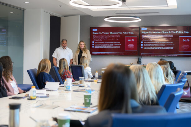 A man and woman stand in front of a large meeting room, presenting to a group of seated people. Two large screens behind them display a presentation titled “Rogue AI, Vendor Chaos & The Policy Gap.”. South Florida Business & Wealth