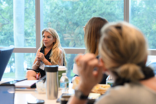 A woman with long blonde hair speaks to two other women during a meeting in a bright conference room with large windows and a view of trees outside. A table with drinks and snacks is in the foreground. South Florida Business & Wealth