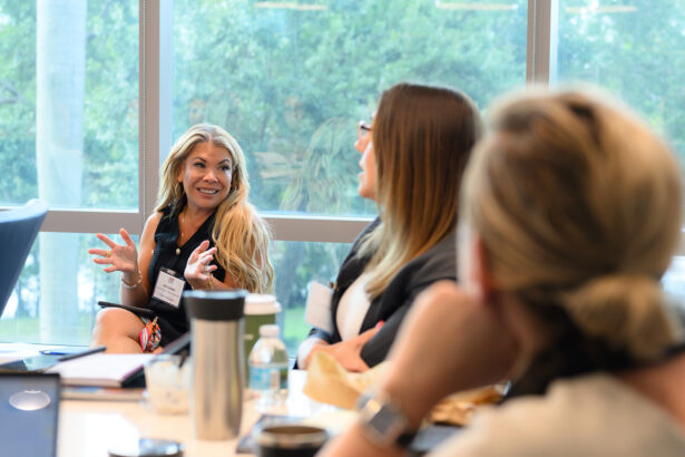 A woman with long blonde hair gestures while speaking to two colleagues at a conference table in a bright meeting room with large windows and a view of trees outside. South Florida Business & Wealth