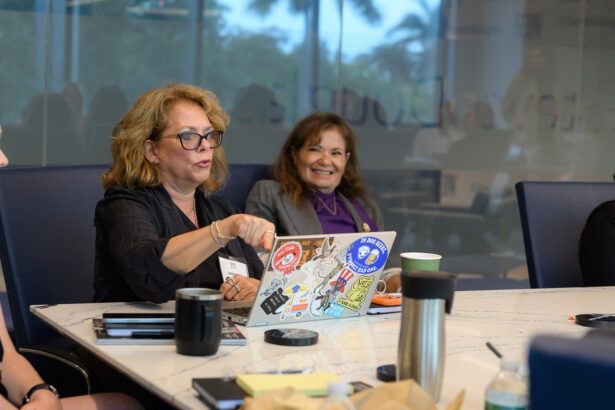 Two women sit at a conference table during a meeting. One gestures while speaking, and both are smiling. Laptops, coffee cups, and notepads are on the table, and the background shows a glass wall with a view of trees. South Florida Business & Wealth
