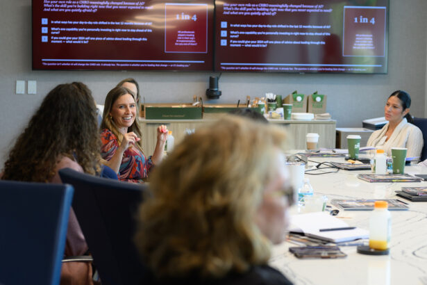A group of people sit around a conference table in a meeting room. A woman in a patterned dress speaks, while others listen. Large screens behind them display statistics and information in red and white text. South Florida Business & Wealth