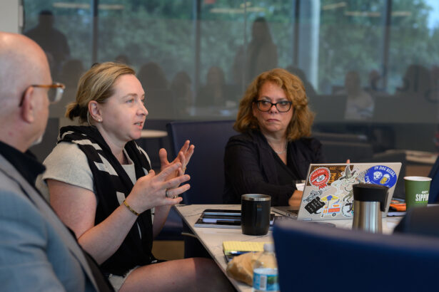A woman gestures while speaking at a conference table with two other people. Laptops, notebooks, drinks, and papers are on the table; a large window shows an outdoor scene in the background. South Florida Business & Wealth