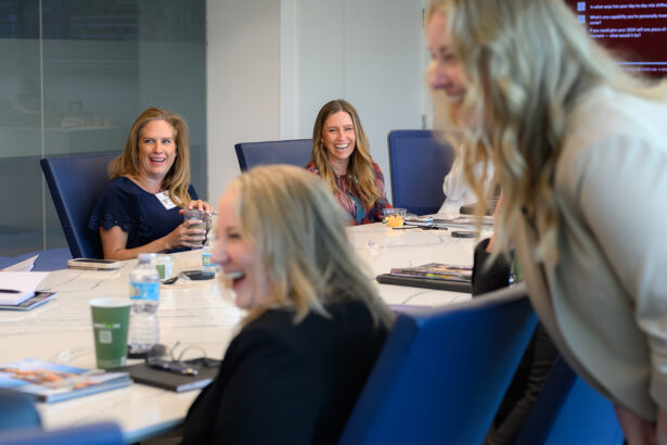 A group of women sit and stand around a conference table, smiling and laughing together during a meeting in a modern office setting. South Florida Business & Wealth