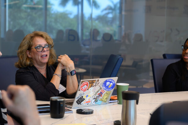 A woman with glasses speaks at a conference table, sitting in front of a laptop covered in colorful stickers. Other people are seated nearby. The room has large windows with a view of trees outside. South Florida Business & Wealth