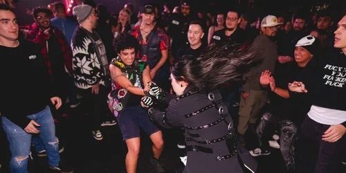 A group of people at a concert or club watch as two individuals in the center energetically dance or playfight, surrounded by cheering and smiling onlookers in casual, colorful clothing. South Florida Business & Wealth