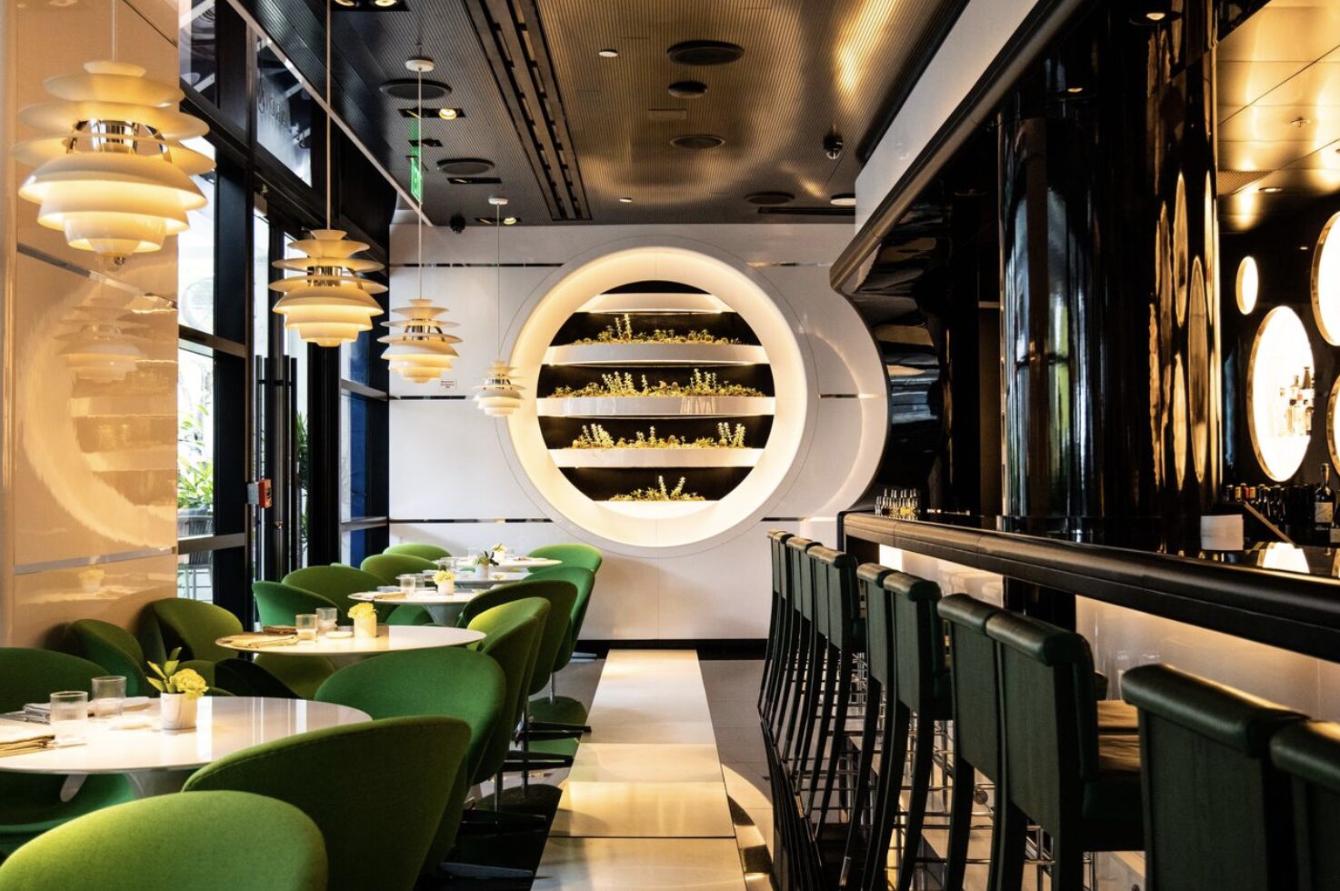 Modern restaurant interior with green chairs, black bar stools, oval shelves with plants, round lights, and a sleek, black-and-white color scheme. Large windows let in natural light. South Florida Business & Wealth