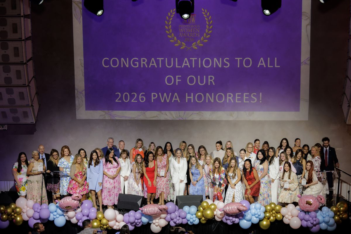 A large group of women stand on stage, smiling and holding awards. Behind them, a purple screen reads, "Congratulations to all of our 2026 PWA Honorees!" Pink and gold balloons decorate the stage. South Florida Business & Wealth