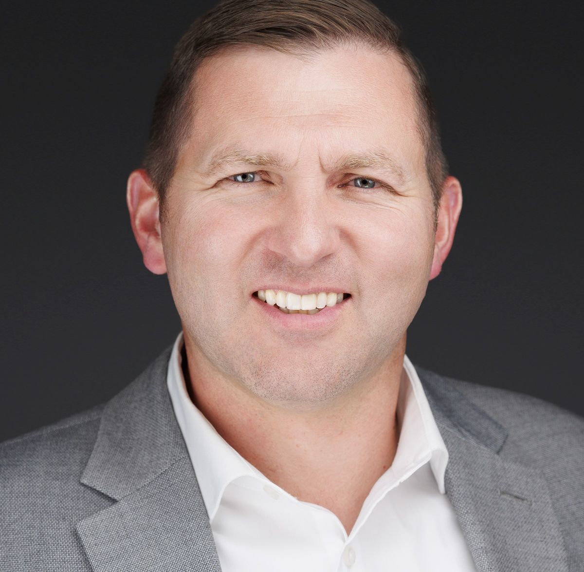 A man with short brown hair wearing a gray suit jacket and white dress shirt smiles at the camera against a dark background. South Florida Business & Wealth