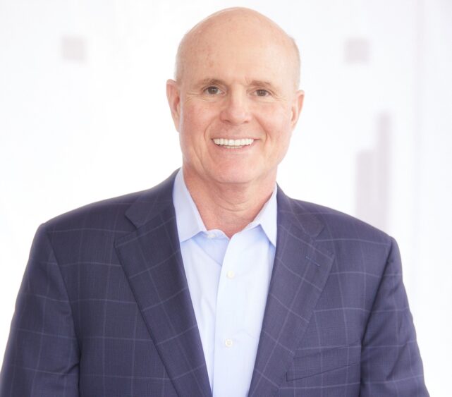 Bald man wearing a blue checked suit jacket and light blue shirt, smiling at the camera, with a bright, blurred white background. South Florida Business & Wealth