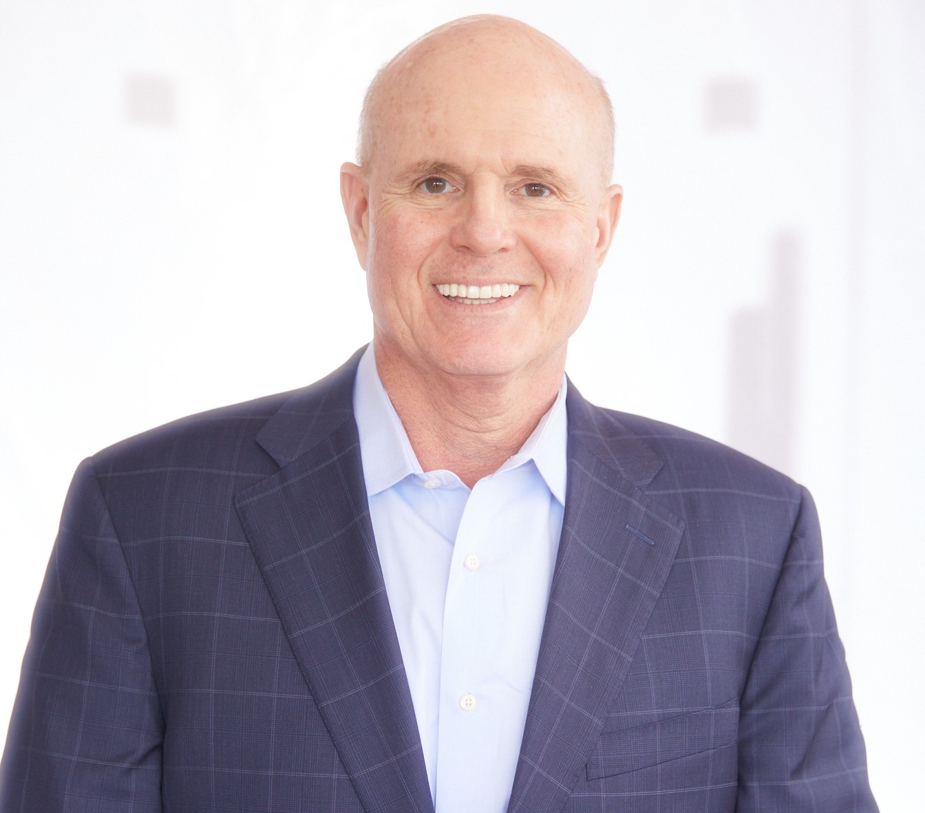 Bald man wearing a blue checked suit jacket and light blue shirt, smiling at the camera, with a bright, blurred white background. South Florida Business & Wealth