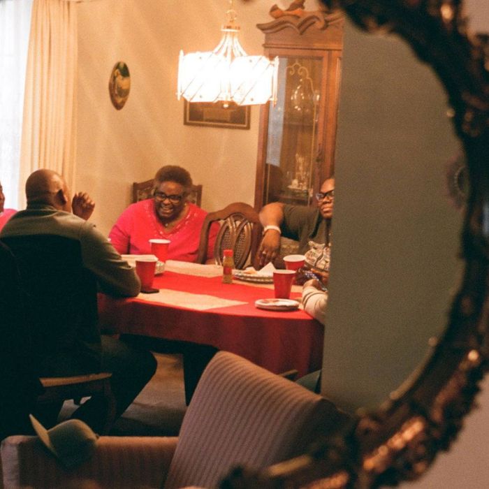 Four people sit around a dining table with a red tablecloth, talking and laughing. The scene is warmly lit and reflected in an ornate mirror, with red cups and plates on the table. South Florida Business & Wealth