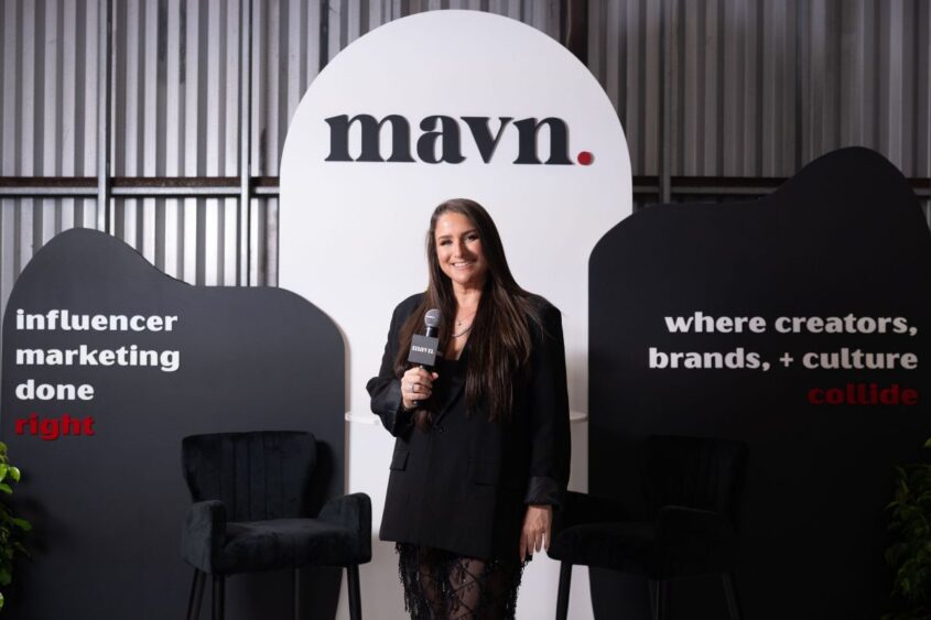 A woman in a black outfit stands holding a microphone in front of a MAVN sign, with two black chairs and display boards reading “influencer marketing done right” and “where creators, brands, + culture collide.”. South Florida Business & Wealth