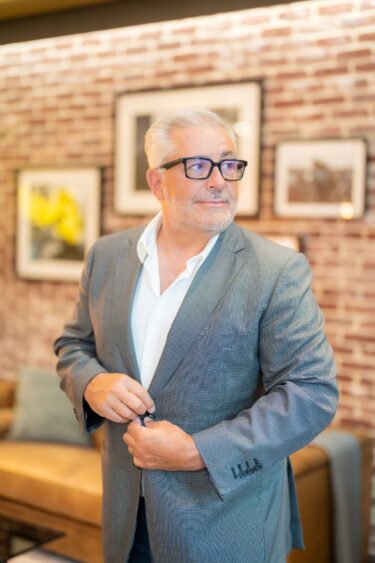A mature man with gray hair and glasses, wearing a gray suit and white shirt, stands indoors and buttons his jacket. There is a brick wall with framed art and a beige couch in the background. South Florida Business & Wealth