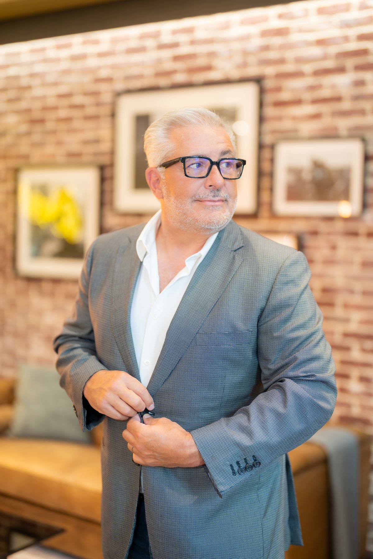 A mature man with gray hair and glasses, wearing a gray suit and white shirt, stands indoors and buttons his jacket. There is a brick wall with framed art and a beige couch in the background. South Florida Business & Wealth