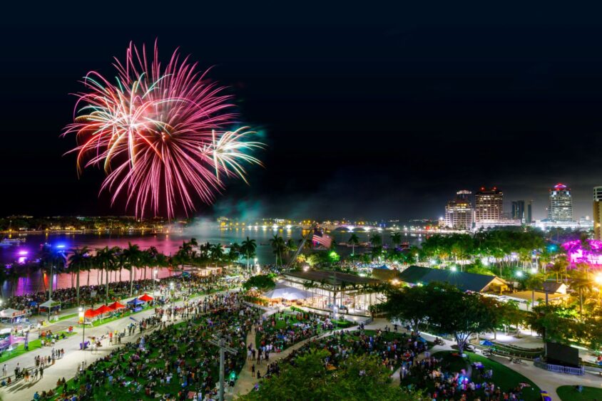 Colorful fireworks light up the night sky over a lively waterfront park filled with people, palm trees, and city buildings illuminated in the background. South Florida Business & Wealth