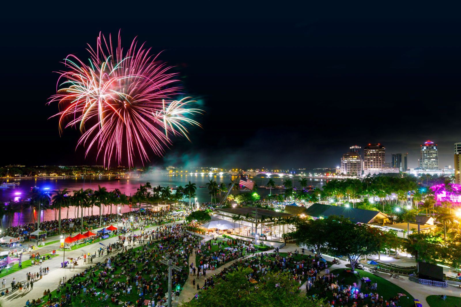 Colorful fireworks light up the night sky over a lively waterfront park filled with people, palm trees, and city buildings illuminated in the background. South Florida Business & Wealth