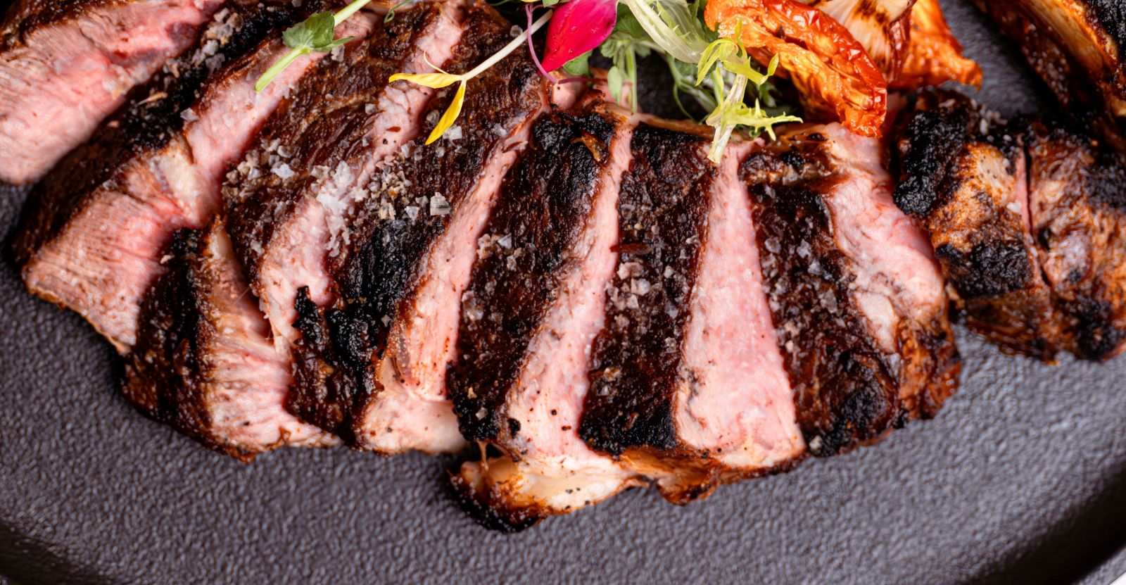 Sliced medium-rare steak with a charred crust, topped with coarse sea salt, served on a dark plate with a garnish of greens and a roasted tomato. South Florida Business & Wealth