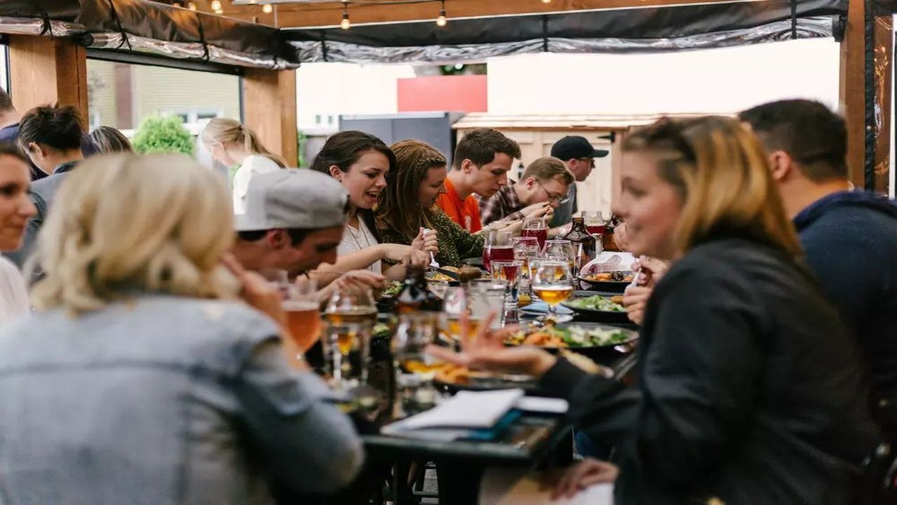 A group of people sit around a long outdoor table, enjoying food and drinks together. The scene appears lively and social, with various dishes, glasses, and conversations taking place under string lights. South Florida Business & Wealth