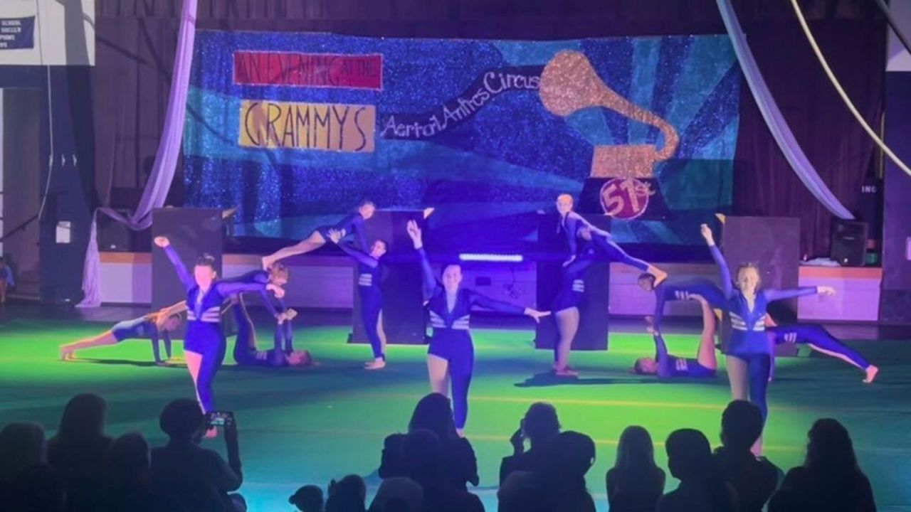 A group of performers in blue costumes pose and balance in a gymnasium under stage lights, with an audience watching. A backdrop reads "GRAMMYS" and "Aerial Artists Circus," featuring a large gold Grammy trophy symbol. South Florida Business & Wealth
