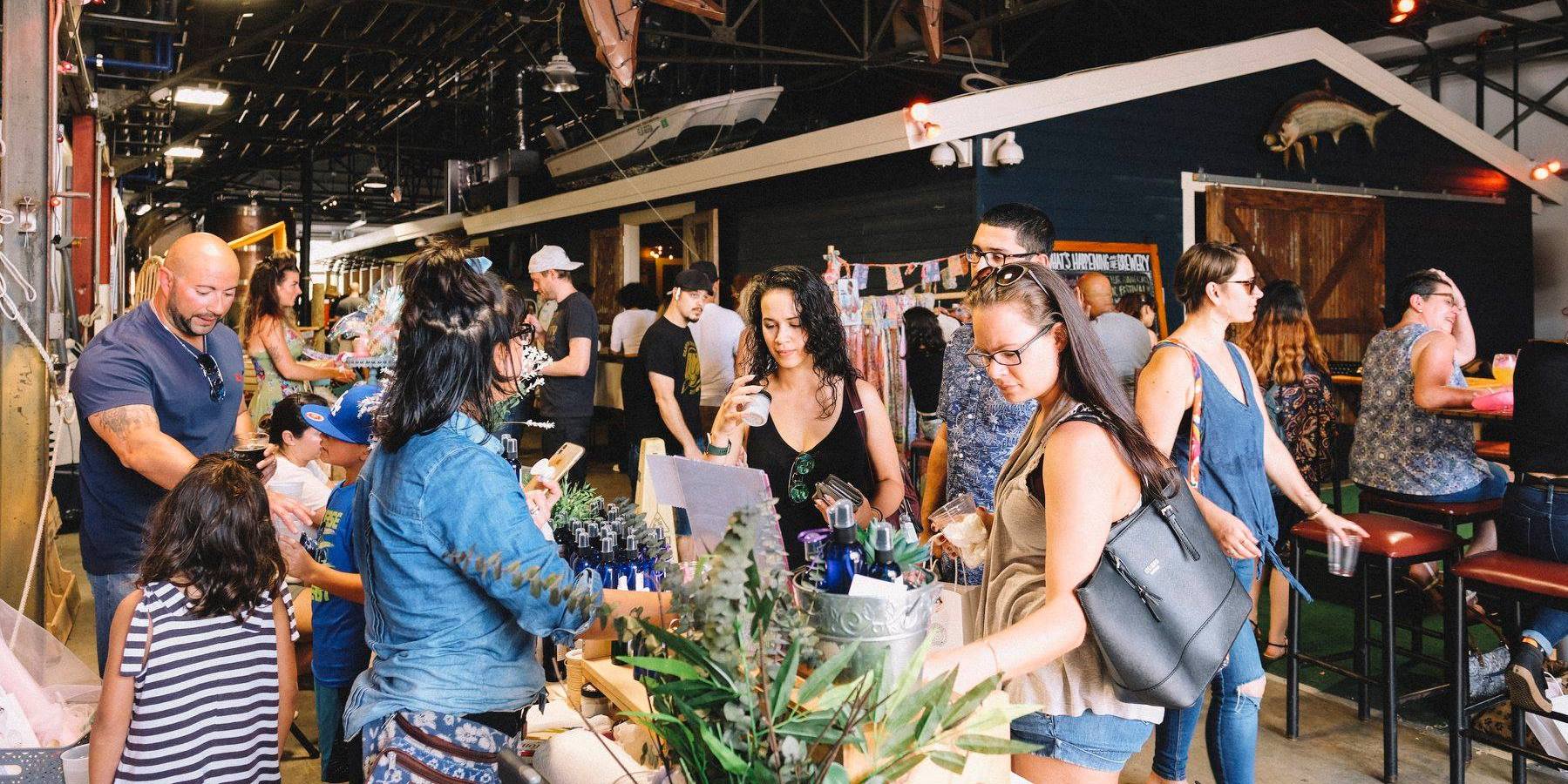 People browse tables with goods at an indoor market or fair. The setting is lively, with adults and children interacting and exploring different booths in a spacious, decorated area. South Florida Business & Wealth