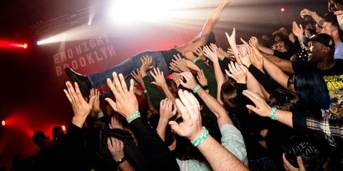 A crowd at a concert lifts a person above their heads for crowd surfing. Many hands are reaching up, and colorful stage lights illuminate the scene. A sign in the background reads "EMO NIGHT BROOKLYN. South Florida Business & Wealth