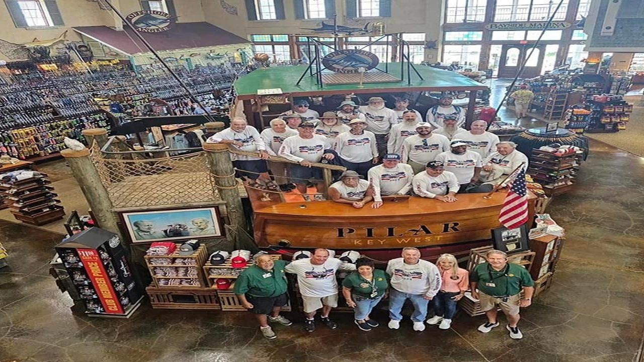 A group of people poses on and around a boat display labeled "PILAR Key West" inside a large store with fishing gear, clothing, and various merchandise visible in the background. South Florida Business & Wealth