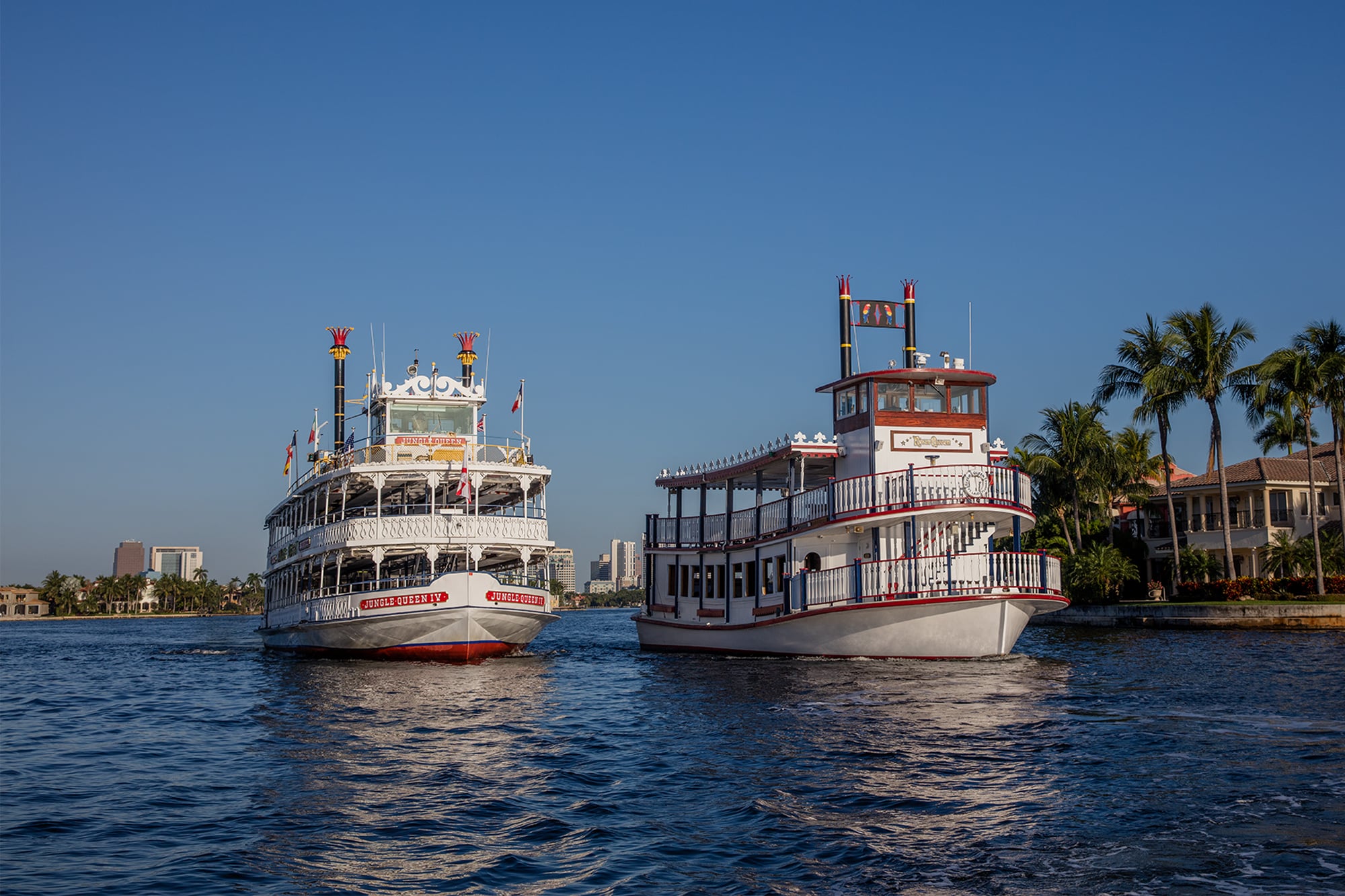 Two white riverboats with red trim and decorative flags sail on a calm waterway, with palm trees, houses, and a clear blue sky in the background. South Florida Business & Wealth