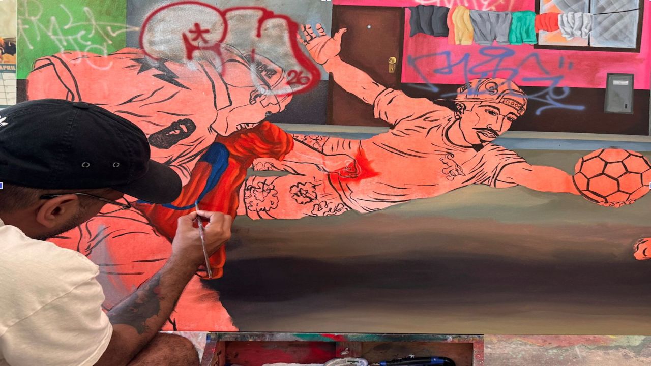 An artist wearing a black cap works on a colorful mural showing two handball players in action, with some graffiti and colorful fabrics in the background. The mural appears partially finished. South Florida Business & Wealth
