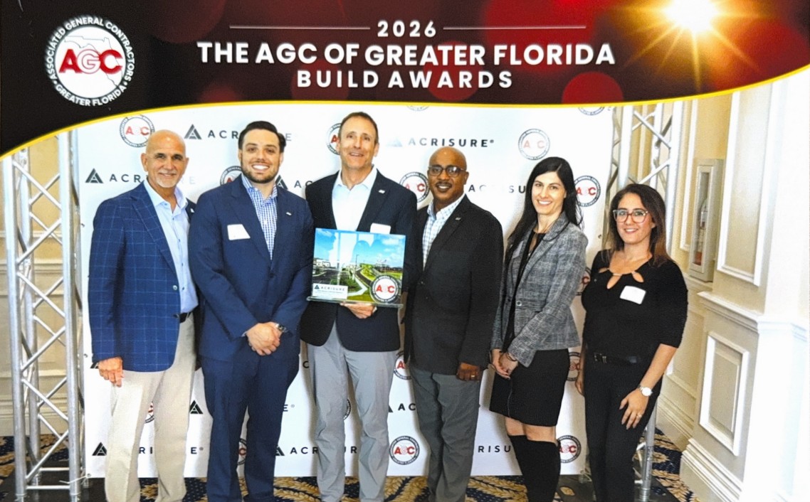 Six professionally dressed people stand together smiling at the 2026 AGC of Greater Florida Build Awards event, in front of a branded backdrop and under bright lights. One man holds a plaque or booklet. South Florida Business & Wealth