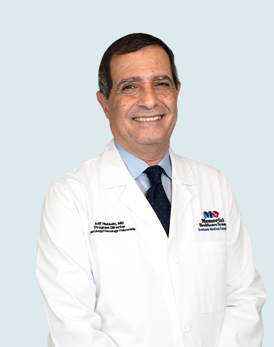 A smiling man in a white doctor’s coat and navy blue tie stands against a light background. The coat has embroidered text and a heart logo on the chest. South Florida Business & Wealth