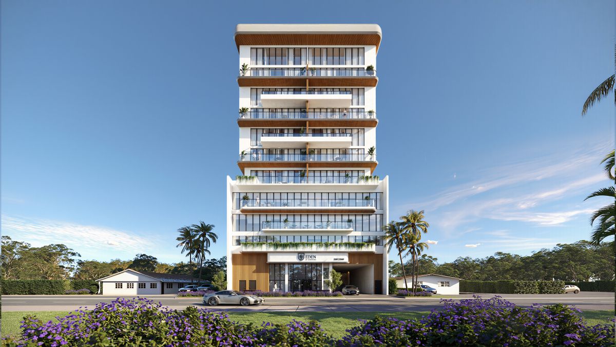A modern multi-story building with large glass balconies, palm trees on both sides, cars parked in front, and purple flowers in the foreground under a clear blue sky. South Florida Business & Wealth
