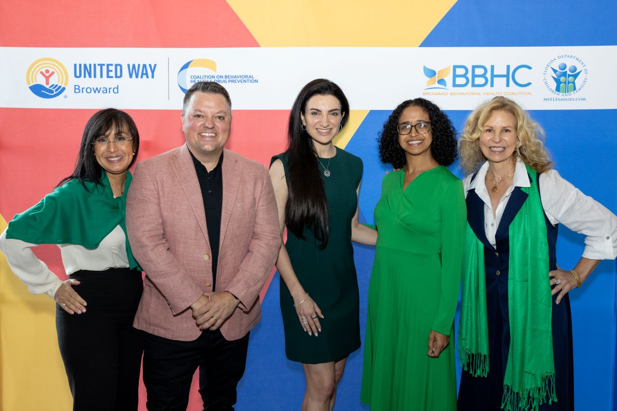 Five people stand smiling in front of a colorful backdrop with logos for United Way Broward, BBHC, and two other organizations. They are dressed in business or semi-formal attire. South Florida Business & Wealth