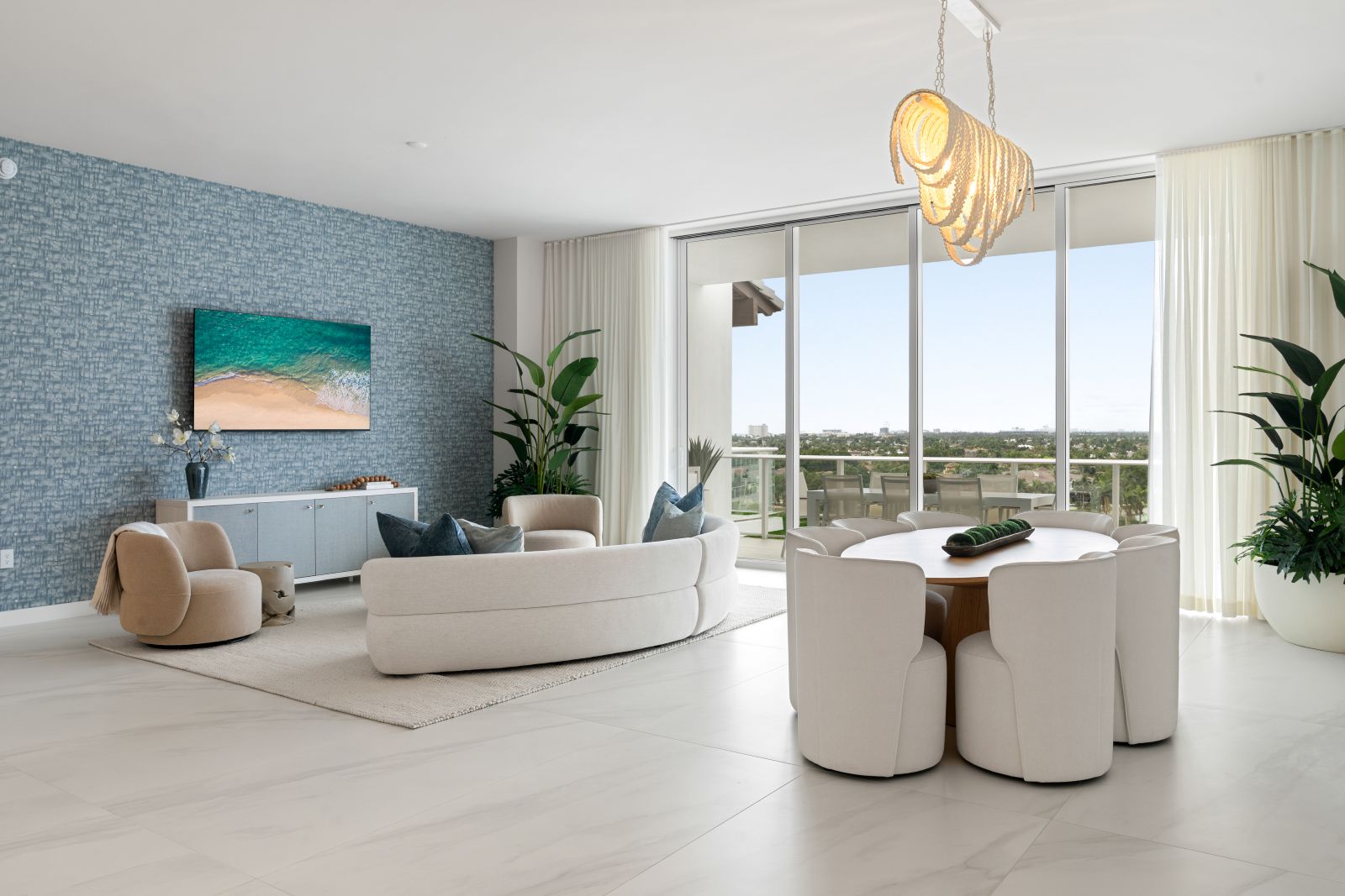 Modern living and dining area with white furniture, large windows, indoor plants, blue accent wall with TV, round dining table, and pendant light. Balcony provides a view of greenery and sky. South Florida Business & Wealth