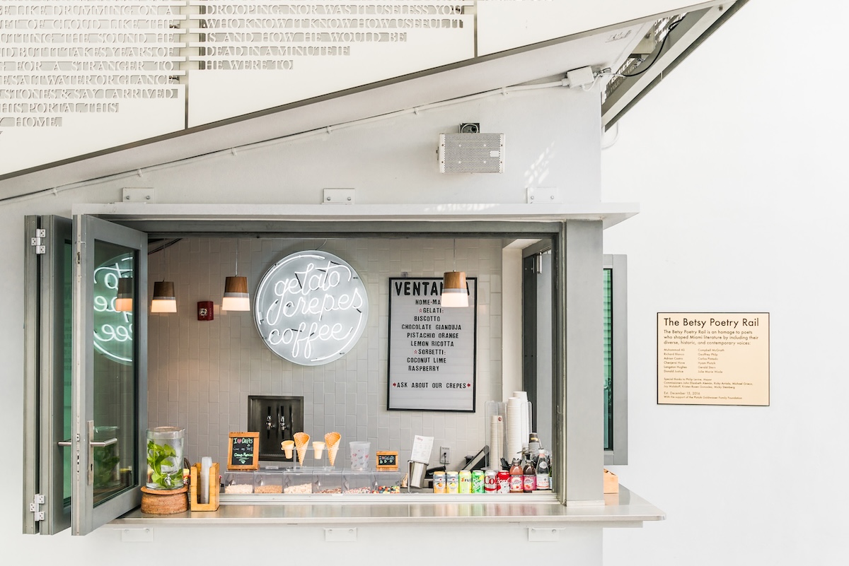 A small modern café counter with open windows, a neon sign reading “gelato crepes coffee,” a menu board, and various snacks and drinks on display. There’s a plaque to the right, and minimal decor. South Florida Business & Wealth