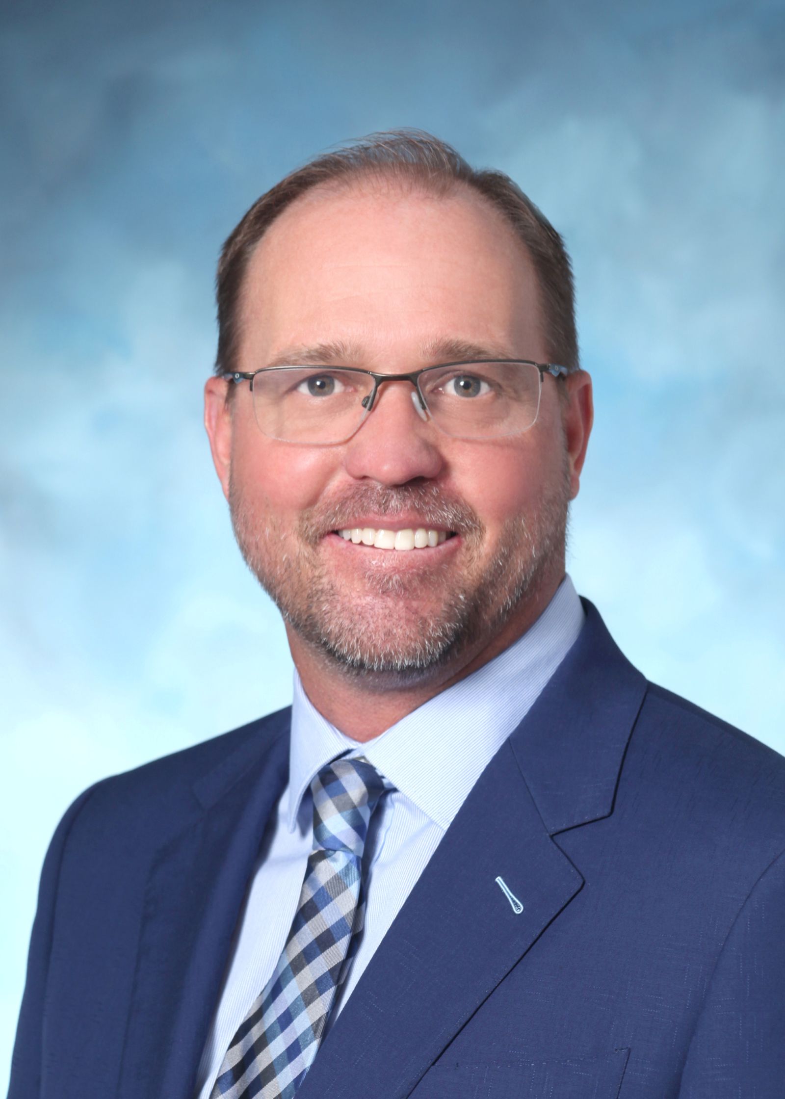 A man with short light brown hair, a trimmed beard, and glasses is wearing a blue suit, a white shirt, and a blue patterned tie. He is smiling and posed against a light blue background. South Florida Business & Wealth