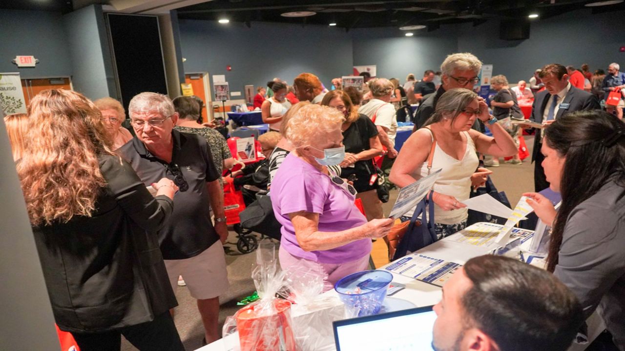 A crowded indoor event where people of various ages are gathered, talking and examining brochures at tables with promotional items and displays. South Florida Business & Wealth